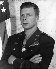 black and white photo of a man in uniform with arms crossed next to an american flag