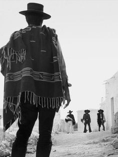 black and white photograph of man in hat with ponchy over his shoulders walking down the street