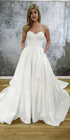 a woman standing in front of a wall wearing a white wedding dress with pockets on the skirt