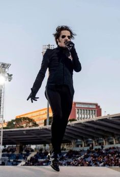 a man in black jacket and sunglasses walking on stage with microphone to his ear while people watch from the bleachers