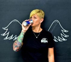 a woman is drinking from a can with wings painted on the wall in the background