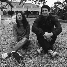 two people sitting on the grass in front of a house and one person is looking at the camera