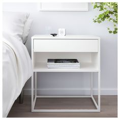 a white bedside table with a magazine on it next to a plant and a bed