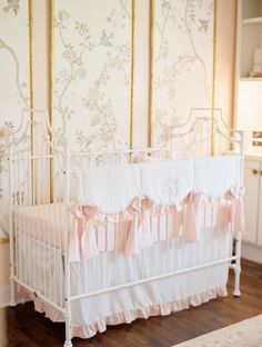 a white crib with pink ruffled bedding and wallpaper in the background