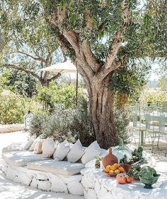 an outdoor seating area under a tree with pillows and fruit on the ground next to it