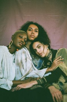 three women sitting next to each other in front of a pink background, one is holding her arm around the other's neck