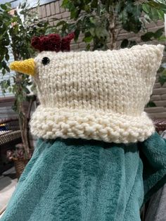 a knitted pillow with a chicken head on it's back, sitting in front of a potted plant