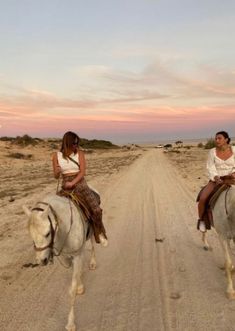 two women riding horses down a dirt road in the desert at sunset or dawn,