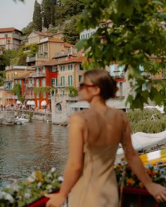 a woman in a dress is standing by the water looking at buildings and boats on the shore