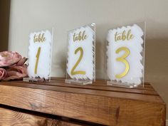 three clear acrylic table numbers sitting on top of a wooden shelf next to pink flowers