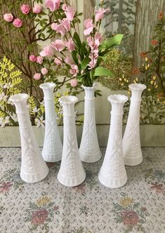 four white vases sitting next to each other on a table with flowers in the background