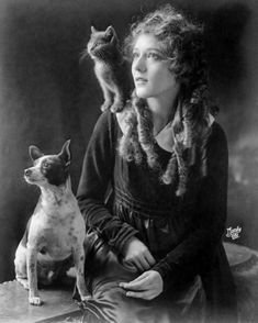 an old black and white photo of a woman with a cat on her shoulders sitting next to a dog