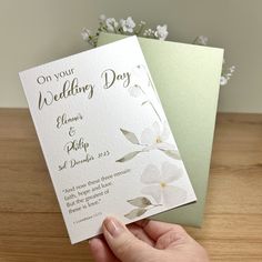 a hand holding two wedding cards on top of a wooden table next to a vase with flowers