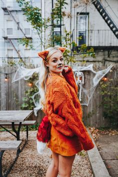 a woman in an orange sweater and red furry ears is posing for the camera with her hands on her hips