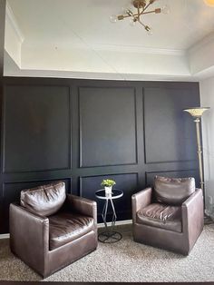 two brown leather chairs sitting next to each other in front of a wall with black paneling