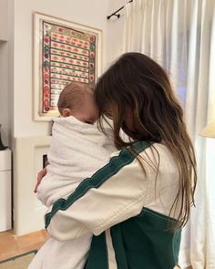 a woman holding a baby wrapped in a white and green blanket while standing next to a window