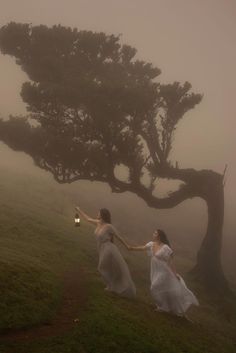 two women in white dresses holding hands and walking up a hill with a tree behind them