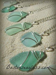 four sea glass necklaces are hanging from silver chains on the sand, and one is green