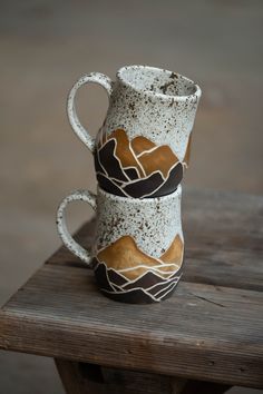 two coffee mugs sitting on top of a wooden table with mountains painted on them