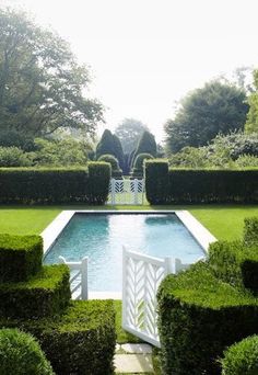 a pool surrounded by hedges and trees in the middle of a garden with a white gate