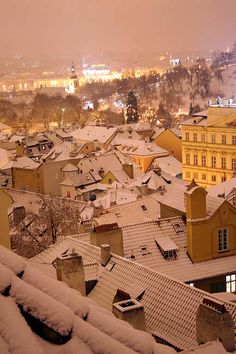 snow covered rooftops and buildings in a city at night with lights on the horizon