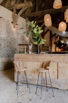 This shows a close up focus on a wedding bar display in a rustic barn. The bar display features cane bar stools, fake plants, wooden crates and vases of dried flowers. Above the bar are cane lampshades with warm light bulbs. Rustic Bar Stools, Rustic Bar