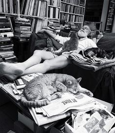 black and white photograph of man laying on couch with cats sleeping next to him in front of bookshelves