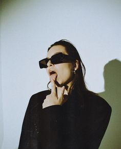 a woman wearing sunglasses and holding her hand up to her mouth while standing in front of a white wall