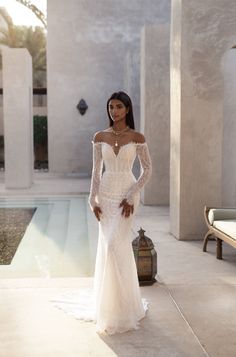 a woman standing in front of a pool wearing a white wedding dress with long sleeves