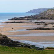 people are walking on the beach near the water and cliffs in the distance, with one person carrying a surfboard