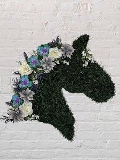 a horse made out of grass with flowers on it's head is shown in front of a white brick wall