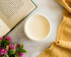 an open book and some pink flowers on a white marble table with a yellow scarf