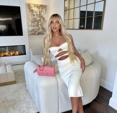 a woman in a white dress sitting on a couch holding a pink purse and posing for the camera