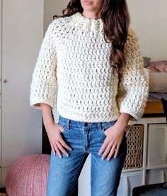 a woman standing in front of a bed wearing blue jeans and a white crochet sweater