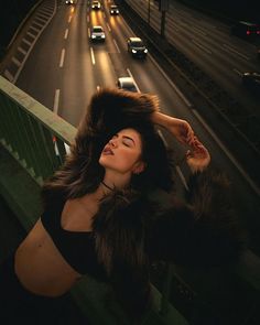 a woman wearing a fur coat standing next to a bridge at night with cars on the road in the background