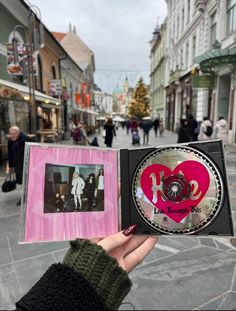 a person holding up a cd in the middle of a street with people walking on it