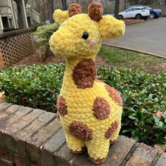 a crocheted giraffe sitting on top of a brick wall next to bushes