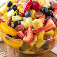 a fruit salad in a glass bowl on top of a wooden table next to a napkin