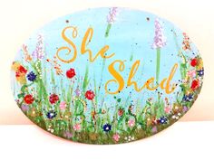 a painted sign that says she shed with flowers and grass in the foreground on a white background