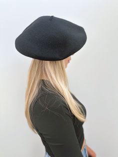 a woman with long blonde hair wearing a black beret and denim jeans, standing in front of a white wall