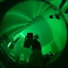 a man taking a photo in front of a green light with his cell phone while standing next to a spiral staircase