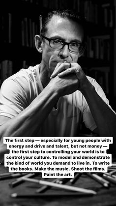 a man with glasses sitting at a table in front of a book shelf and holding his hands to his mouth