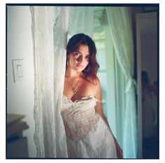 a woman in a white dress is standing behind a curtain and looking at the camera