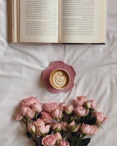 a cup of coffee and some pink roses on a white bed with an open book