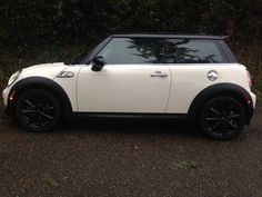 a small white car parked in front of some bushes and trees with black rims