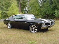 an old black muscle car parked in the middle of a field with trees behind it