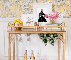 a gold bar cart with wine glasses and bottles on it in front of a floral wallpaper