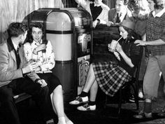 a black and white photo of people sitting around an old fashioned soda dispenser