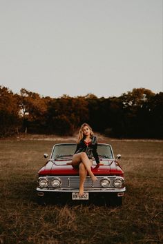 a woman sitting on the hood of a red car in a field with trees behind her