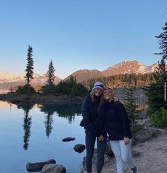 two women standing next to each other near a body of water with mountains in the background